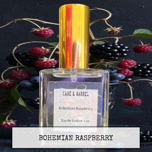 May include: A clear glass perfume bottle with a gold cap, labeled "Cake & Barrel Bohemian Raspberry Eau De Parfum 1 oz." The bottle is surrounded by fresh raspberries, blackberries, and blueberries against a dark background. The text "Bohemian Raspberry" is at the bottom.