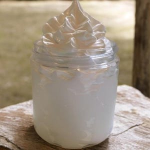 May include: A clear glass jar filled with white cream, topped with a swirl of whipped cream. The jar sits on a weathered wooden surface, with a blurred background of greenery.