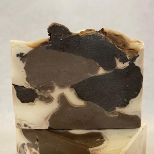 May include: Close-up of a handmade soap bar with a marbled design. The soap features a blend of colors, including white, light brown, dark brown, and black, creating a unique, natural pattern. The top of the bar has a textured, uneven surface.