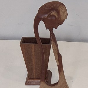 May include: A wooden sculpture of a person with their head bent down, holding a wooden vase. The vase is rectangular and has a square base. The sculpture is made of dark brown wood.