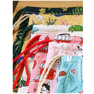 May include: A stack of five colourful fabric drawstring bags with various patterns. The top bag is green with a floral design, the second is yellow with a green leaf pattern, the third is beige with a car design, the fourth is pink with a red and white pattern, and the bottom bag is blue with a green pine tree design.