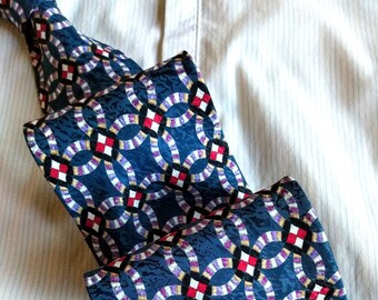 May include: A blue patterned tie with white, red, and purple circles and squares. The tie is folded and laid on a white striped shirt.