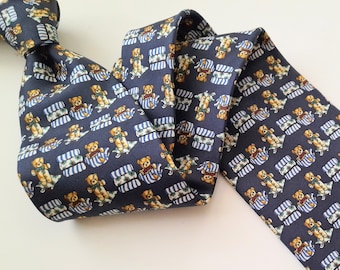 Könnte beinhalten: Eine dunkelblaue Seidenkrawatte mit einem sich wiederholenden Muster von Teddybären, die blau-weiß gestreifte Pyjamas tragen. Die Bären halten Gießkannen und sitzen auf einem blau-weiß gestreiften Hintergrund.