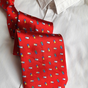 May include: A red silk tie with a pattern of small, colourful rectangles. The tie is folded and laid on a white, striped shirt.