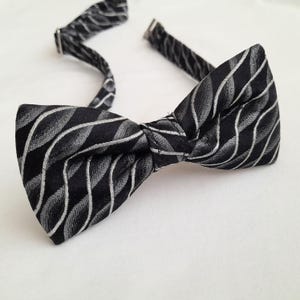 Puede incluir: Una pajarita negra con un patrón de ondas plateadas repetidas. La pajarita preatada tiene una correa ajustable para el cuello con herrajes metálicos plateados. La tela tiene un ligero brillo, sobre un fondo blanco.