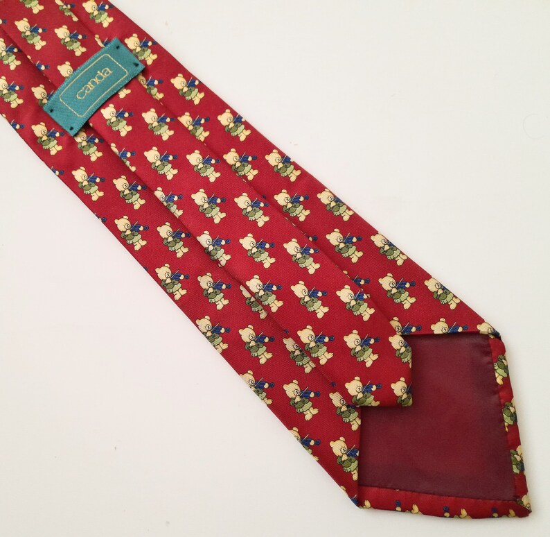 May include: Red silk tie with a pattern of small teddy bears playing musical instruments. The tie has a burgundy lining and a green label that says "canda".