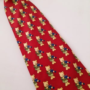 May include: A red silk tie with a repeating pattern of teddy bears playing the violin. The bears are yellow with brown noses and blue eyes. The tie has a dark blue trim.
