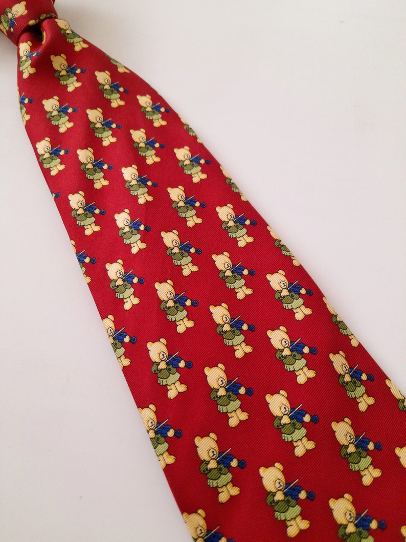 May include: A red silk tie with a repeating pattern of teddy bears playing the violin. The bears are yellow with brown noses and blue eyes. The tie has a dark blue trim.