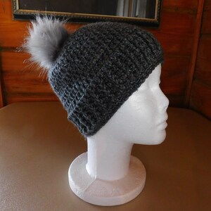 May include: A gray crocheted beanie with a fluffy gray pom pom on top. The beanie is displayed on a white mannequin head.