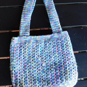 May include: A handmade crochet tote bag in shades of blue, purple, and green. The bag features a textured stitch pattern and sturdy handles. This versatile bag is perfect for carrying everyday essentials.