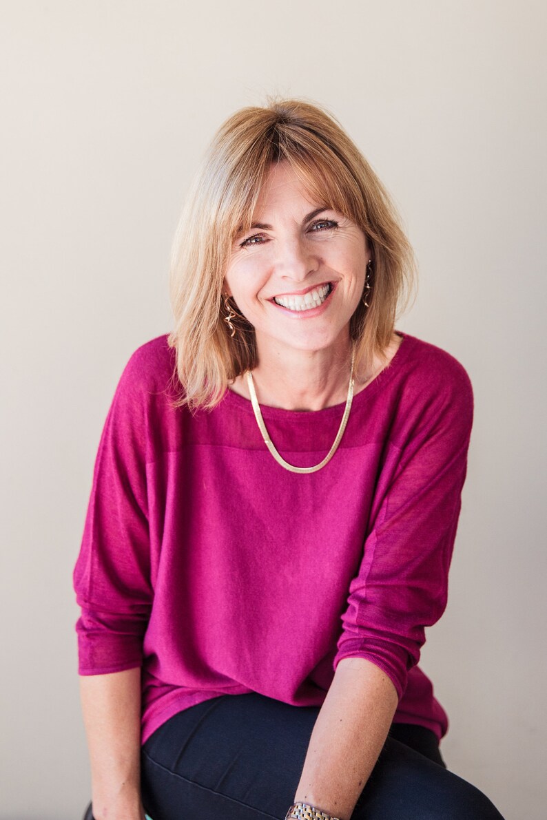 May include: A woman with blonde hair is smiling and wearing a pink sweater and black pants. She is wearing a gold necklace and earrings.
