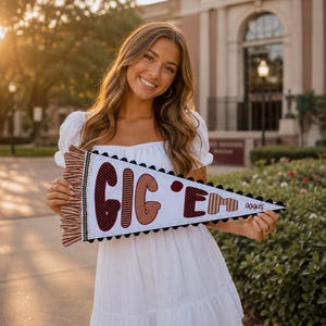 May include: A white triangular felt pennant featuring the maroon text GIG 'EM and the word Aggies in a smaller script font. The pennant is decorated with a maroon and white striped ruffle on the left side and black pom-pom trim along the edges.