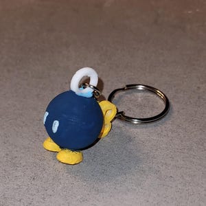 Peut inclure: Porte-clés artisanal représentant une bombe bleue avec des détails blancs et des pieds jaunes. Le porte-clés est équipé d'un anneau argenté. La bombe a une mèche blanche et est un accessoire amusant.