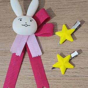 以下が含まれることがあります： ピンクのリボンとストリーマーが付いた白いウサギ型のヘアアクセサリーと、銀色の金属クリップが付いた2つの黄色の星型のヘアクリップ。木製の表面にあります。これらはヘアアクセサリーです。