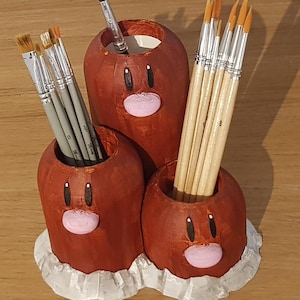 以下が含まれることがあります： 手作りの茶色の木製デスクオーガナイザー。漫画のキャラクターの形をしており、筆を入れる3つの円筒形のコンパートメントがあります。オーガナイザーは白のフリルベースで、黒い目とピンクの口が描かれています。