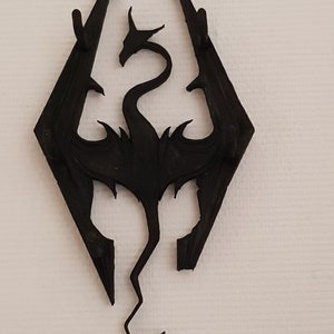 May include: Black 3D printed wall hook shaped like the dragon symbol from the video game The Elder Scrolls V: Skyrim. The hook has five prongs for hanging items.