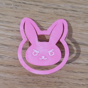 May include: A pink bunny-shaped cookie cutter with a detailed face and ears. The bunny has a stern expression with angled eyebrows and a small nose and mouth. The cutter is made of a solid pink material.