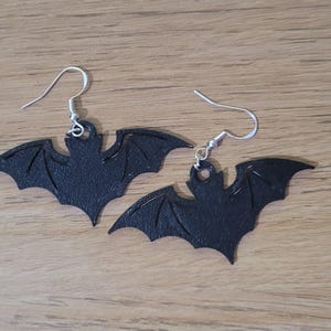 Peut inclure: Une paire de boucles d'oreilles en forme de chauve-souris noire avec une surface texturée et des découpes détaillées sur les ailes. Chaque boucle est fixée à un crochet en métal argenté, conçu pour être suspendu au lobe de l'oreille.