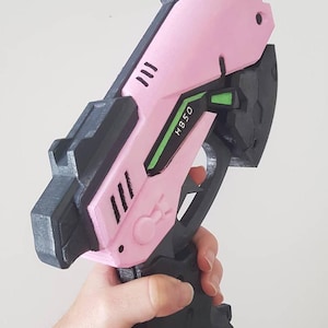 May include: A pink and black toy gun with the text "HB50" in green. The gun has a futuristic design with angled edges and a trigger. The gun is held in a hand.