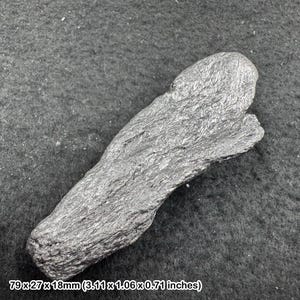 May include: A dark gray, metallic-looking mineral specimen with a rough, fibrous, and crystalline texture. The object has an elongated, irregular shape and measures 3.11 inches by 1.06 inches by 0.71 inches as indicated by the printed text.