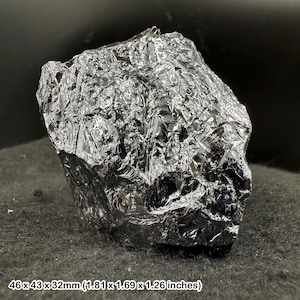 Peut inclure: Spécimen minéral sombre et réfléchissant, de forme irrégulière. La roche est d'un noir profond et brillant avec une surface texturée. Dimensions : 4,6 x 4,3 x 3,2 cm. Le spécimen est présenté sur une surface sombre.
