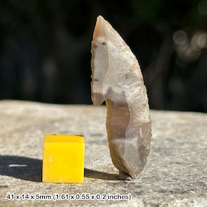 May include: A hand-crafted, light brown stone arrowhead with a unique shape, next to a small, bright yellow cube. The arrowhead measures 1.61 x 0.55 x 0.2 inches. The items are displayed on a stone surface.