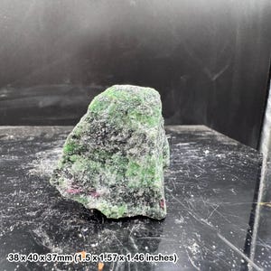 May include: A natural ruby in zoisite specimen, rough-cut. The stone is a mix of green zoisite with ruby inclusions, measuring 38 x 40 x 37mm. The surface is textured, with visible crystal formations.