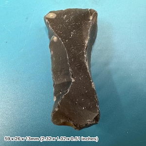May include: A dark gray, roughly shaped stone with a textured surface. The stone measures 2.32 x 1.02 x 0.51 inches. The object is set against a blue background. The stone appears to be a natural, possibly hand-worked item.