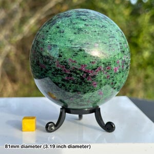May include: A polished sphere of green zoisite with ruby inclusions, 81mm in diameter, is displayed on a black metal stand. The sphere's surface is a mix of green and black, with small red ruby flecks. A small yellow cube is next to the sphere.