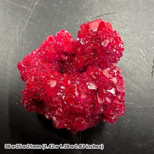 May include: A vibrant red crystal cluster with a jagged, multifaceted surface texture. The specimen measures 36 x 35 x 21 mm, which is equivalent to 1.42 x 1.38 x 0.83 inches. The structure features numerous small, sharp, reflective points.
