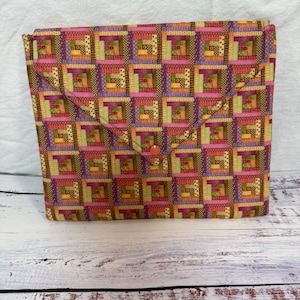 May include: A fabric clutch bag with a geometric patchwork pattern in pink, purple, green, and orange. The bag has a flap closure secured by a single orange button. The background is a white wooden surface.