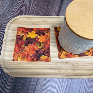 May include: Two square fabric coasters featuring a vibrant autumn leaf pattern in shades of red, orange, and yellow. The coasters are placed on a light-coloured bamboo tray, with one coaster partially covered by a white ceramic jar with a wooden lid.