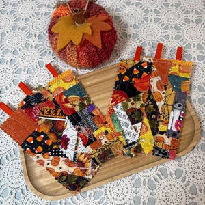 Peut inclure: Deux maniques rectangulaires en tissu matelassé avec un patchwork de motifs sur le thème d'Halloween, incluant des citrouilles, des bonbons et des toiles d'araignée, avec des boucles de suspension rouges. Une citrouille crochetée avec des feuilles en feutrine est présente.