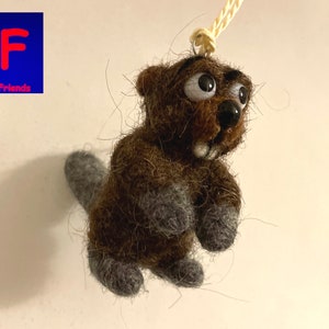 Könnte beinhalten: Ein brauner Filz-Biber mit großen Augen und einem grinsenden Mund. Der Biber hängt an einer Schnur und hat einen grauen Filzschwanz.
