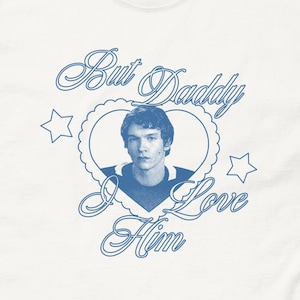 Può includere: T-shirt bianco con grafica blu. La grafica presenta un ritratto all'interno di un cuore, con la scritta "But Daddy I Love Him" e stelle. La maglietta è realizzata in materiale morbido.