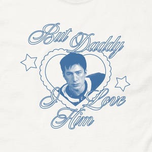 Può includere: T-shirt bianco con grafica blu. La grafica presenta un ritratto all'interno di una cornice a forma di cuore, con la scritta "But Daddy I Love Him" e stelle. La maglietta è realizzata in un materiale morbido e confortevole.