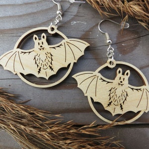 Puede incluir: Un par de pendientes de madera con un diseño de murciélago. Los murciélagos están cortados en madera y tienen un acabado de madera natural. Los pendientes están unidos a ganchos de plata.