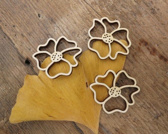 Puede incluir: Tres recortes de flores de madera en color marrón claro. Los recortes tienen forma de flor con cinco pétalos y un círculo central. Los recortes están dispuestos sobre una hoja amarilla.