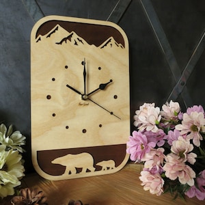 May include: A wooden clock with a mountain scene and two polar bears at the bottom. The clock face is light brown with black hands and a dark brown border. The clock is on a wooden surface.