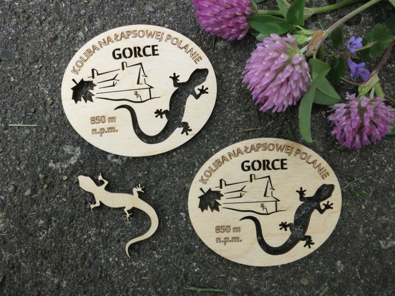 May include: Two wooden signs with the text "KOLIBA NA ŁAPSOWEJ POLANIE" and "GORCE" in Polish. The signs feature a house, a lizard, and the text "850 m n.p.m.".