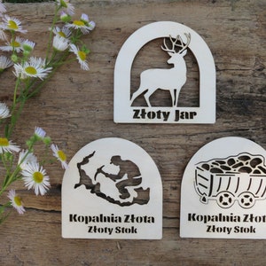 May include: Three wooden plaques with a white background and black cutouts. The top plaque features a deer silhouette and the text "Złoty Jar". The middle plaque features a silhouette of a person mining and the text "Kopalnia Złota Złoty Stok". The bottom plaque features a silhouette of a mine cart and the text "Kopalnia Złota Złoty Stok".