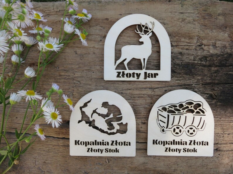 May include: Three wooden plaques with a white background and black cutouts. The top plaque features a deer silhouette and the text "Złoty Jar". The middle plaque features a silhouette of a person mining and the text "Kopalnia Złota Złoty Stok". The bottom plaque features a silhouette of a mine cart and the text "Kopalnia Złota Złoty Stok".