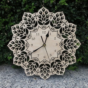 Op de afbeelding: Een houten klok met een mandala-ontwerp en zwarte wijzers. De wijzerplaat van de klok heeft zwarte cijfers van 1 tot en met 12.