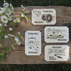 May include: Four wooden signs with laser-cut designs depicting different aspects of the Kłodzko Fortress in Poland. The signs feature the text "Twierdza Kłodzko" and "Most Gotycki" in Polish.