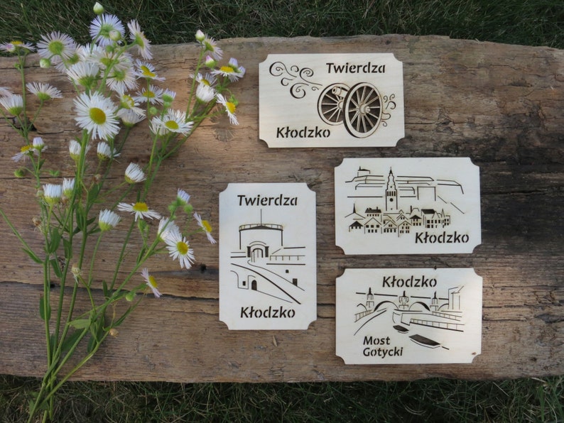 May include: Four wooden signs with laser-cut designs depicting different aspects of the Kłodzko Fortress in Poland. The signs feature the text "Twierdza Kłodzko" and "Most Gotycki" in Polish.