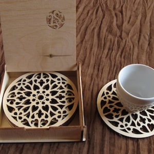 May include: A set of laser-cut wooden coasters with a decorative floral design, presented in a matching wooden box. One coaster supports a white teacup. The coasters and box are made of light-colored wood.