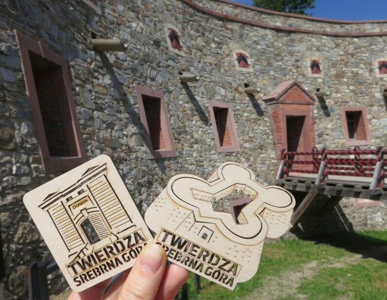 May include: Two wooden souvenirs with the text "TWIERDZA SREBRNA G&Oacute;RA" on them. The souvenirs are in front of a stone building with a red brick doorway.