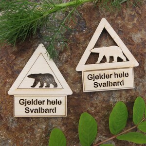 May include: Two wooden triangular warning signs with a silhouette of a polar bear and the text "Gjelder hele Svalbard" which translates to "Applies to all of Svalbard".