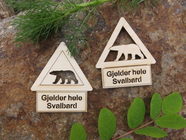 May include: Two wooden triangular warning signs with a silhouette of a polar bear and the text "Gjelder hele Svalbard" which translates to "Applies to all of Svalbard".