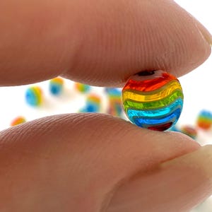 May include: A vibrant, spherical glass bead held between fingers, showcasing a rainbow of colours. The bead's design includes bands of red, orange, yellow, green, blue, and purple. Other beads are visible in the background, slightly out of focus.